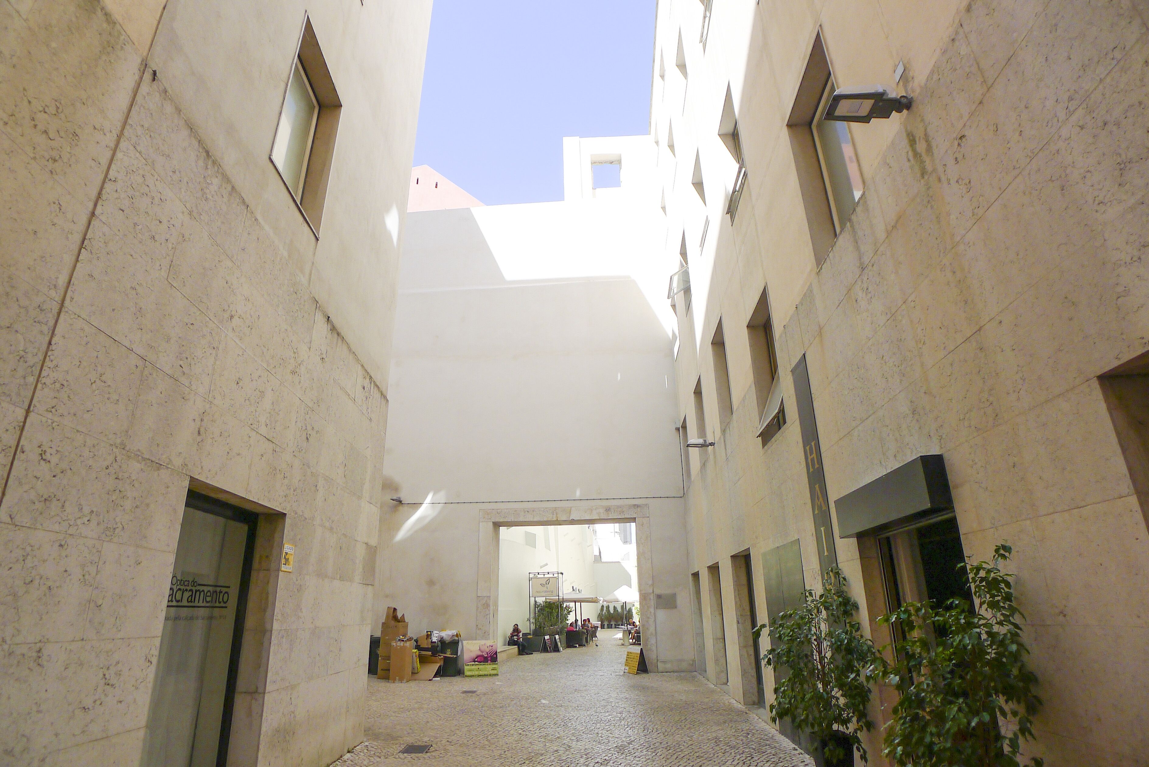 A narrow urban alleyway with a cobblestone floor, flanked by buildings with large windows and a clear blue sky above.