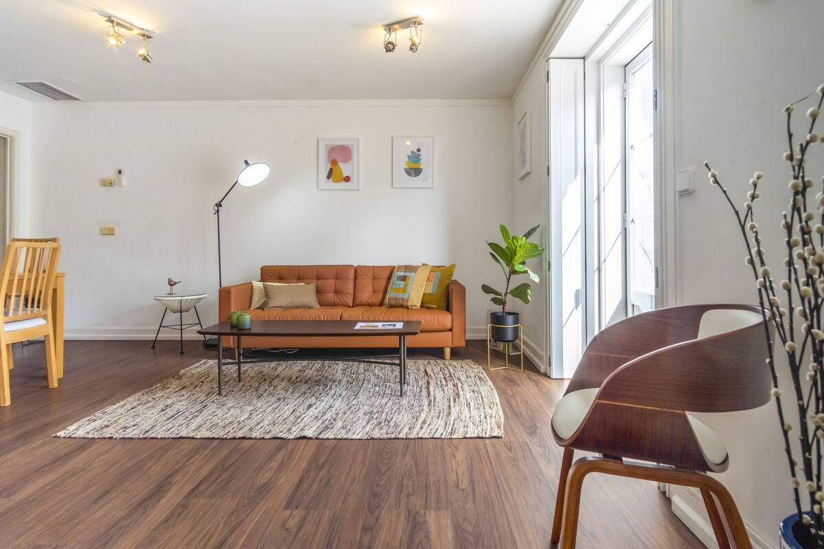 A modern living room with a brown leather sofa, a wooden chair, a coffee table, and a floor lamp. There are two wall-mounted light fixtures and a window with a view of the outside.