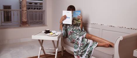 Una persona leyendo un libro en un sofá blanco en una habitación con balcón, una mesita con un plato y una taza, y un pilar decorativo.