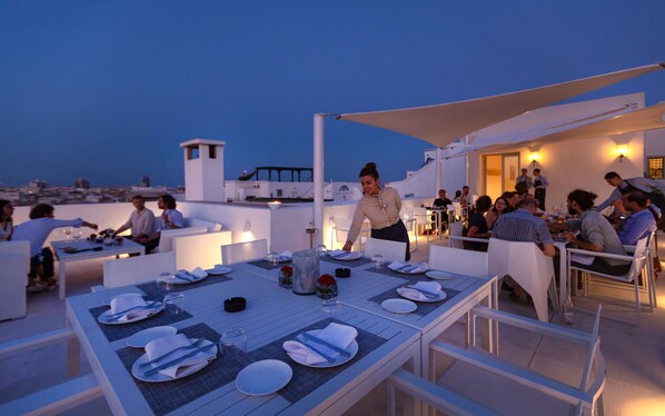 Un espace de restauration en plein air avec des tables dressées pour les clients, un serveur qui s’occupe des clients, et une vue sur la ville en arrière-plan.