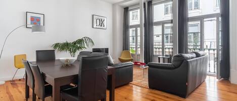 En moderne stue med spisebord, stole, sofa og et stort vindue med gardiner.