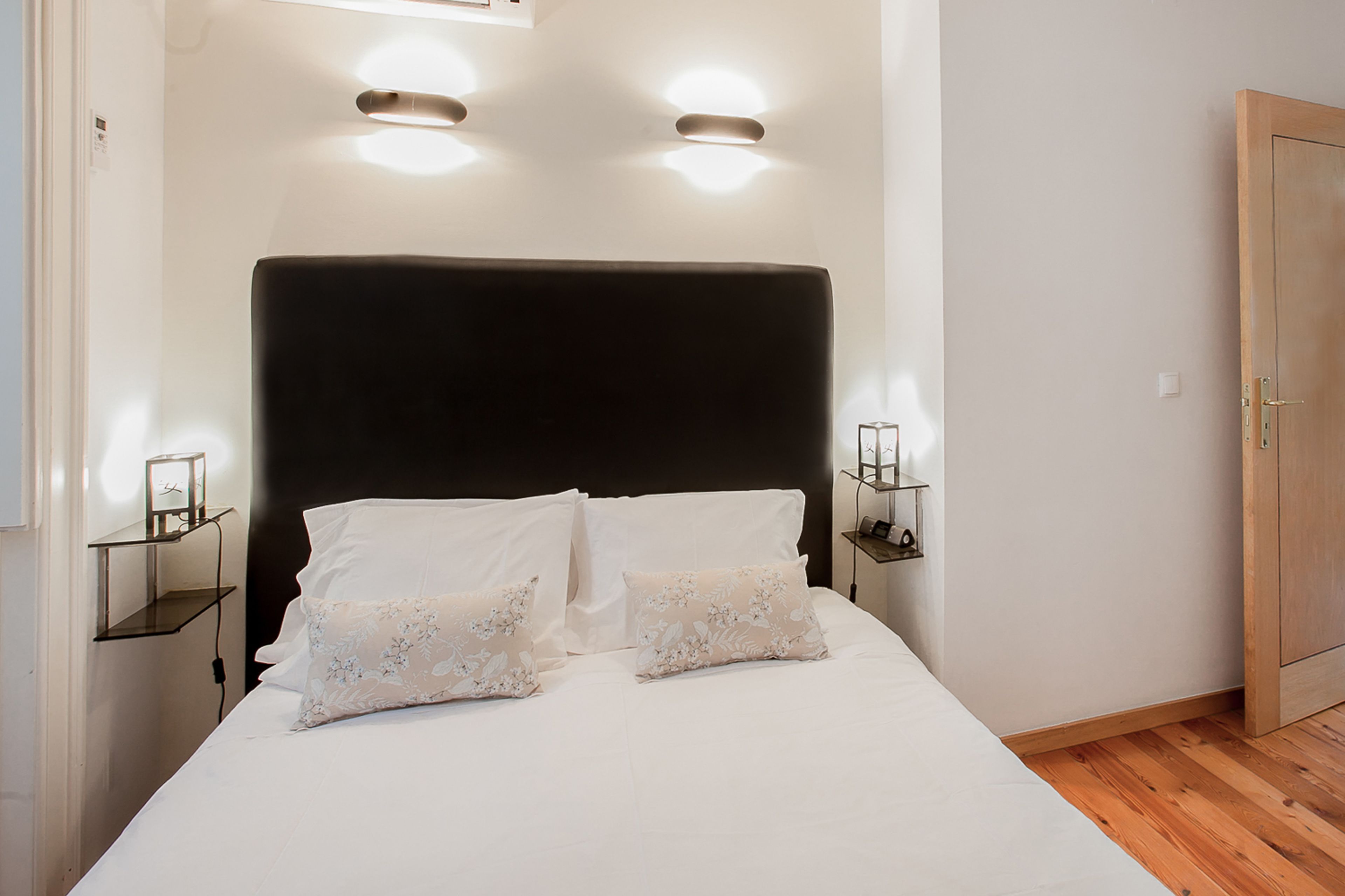 Una cama bien hecha con sábanas blancas y dos almohadones decorativos, con mesitas de noche a cada lado y lámparas fijadas a la pared.