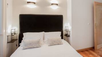 Una cama bien hecha con sábanas blancas y dos almohadones decorativos, con mesitas de noche a cada lado y lámparas fijadas a la pared.