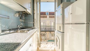 Une cuisine étroite équipée d’un lave-linge encastré, d’un évier et offrant une vue sur un balcon doté d’un sol carrelé et d’une balustrade.