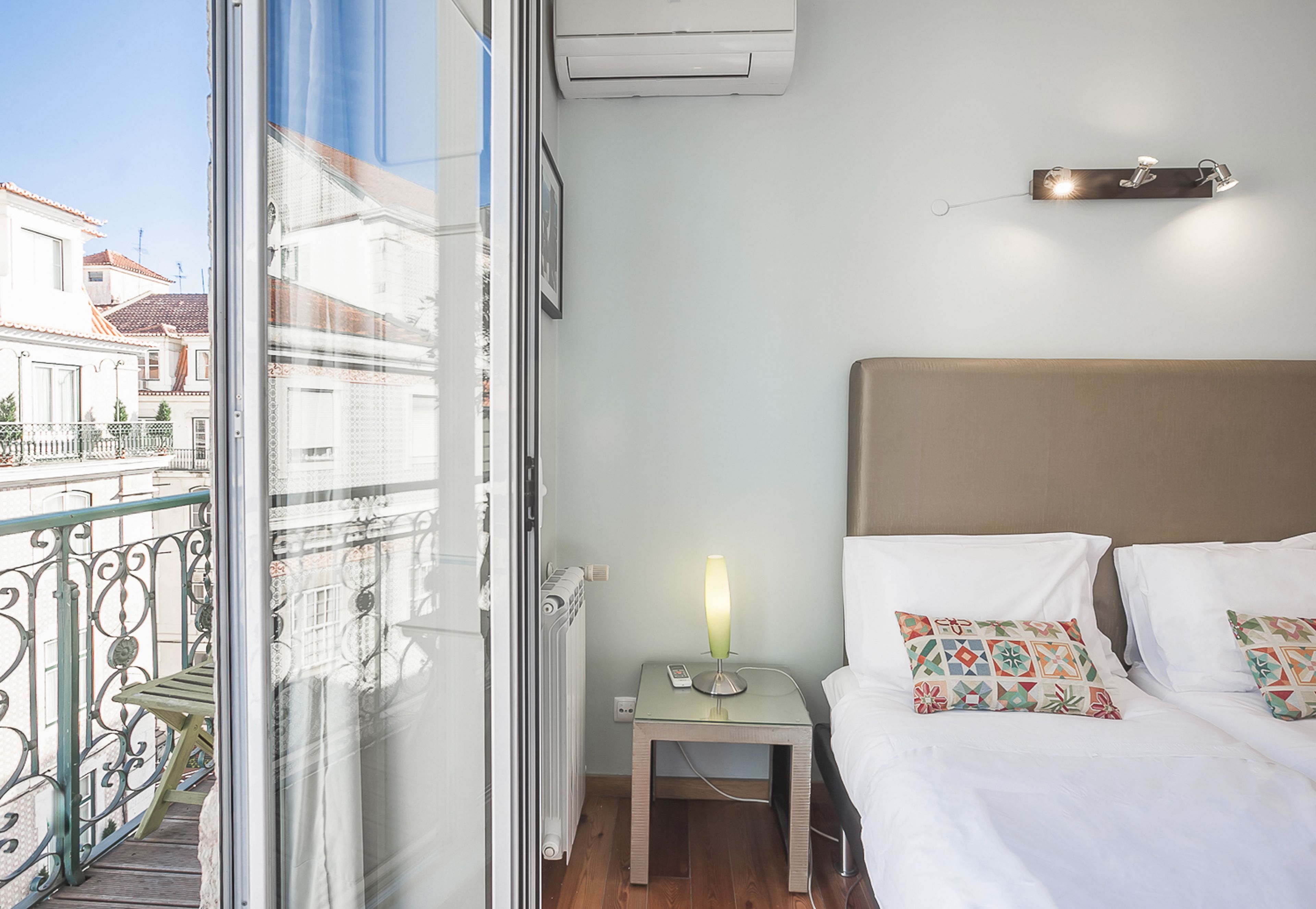 Ein Hotelzimmer mit Balkon, einem Bett mit gemusterten Kissen, einem Nachttisch mit Lampe und einer Wandleuchte.