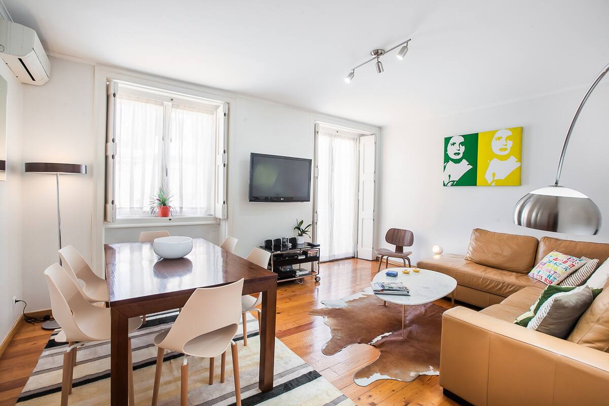 A modern living room with a wooden dining table, white chairs, a brown leather sofa, a white coffee table, and a flat-screen TV.