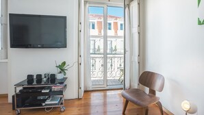 Un salon équipé d’un téléviseur à écran plat, d’une chaise en bois, d’une petite table avec une plante et d’une porte-fenêtre coulissante donnant sur un balcon.