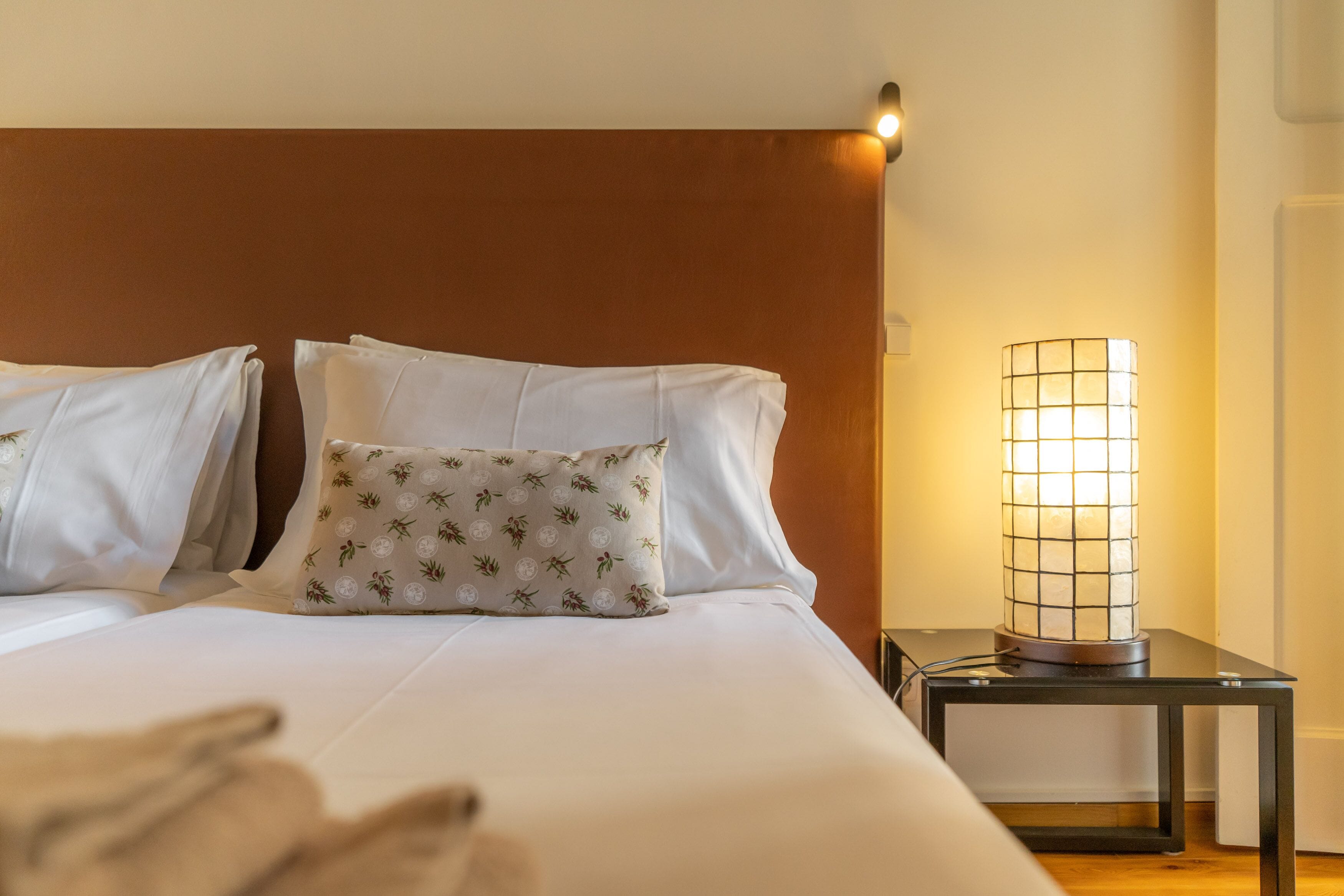 A neatly made bed with white linens and a decorative pillow, a bedside table with a lamp, and a wall-mounted light fixture.
