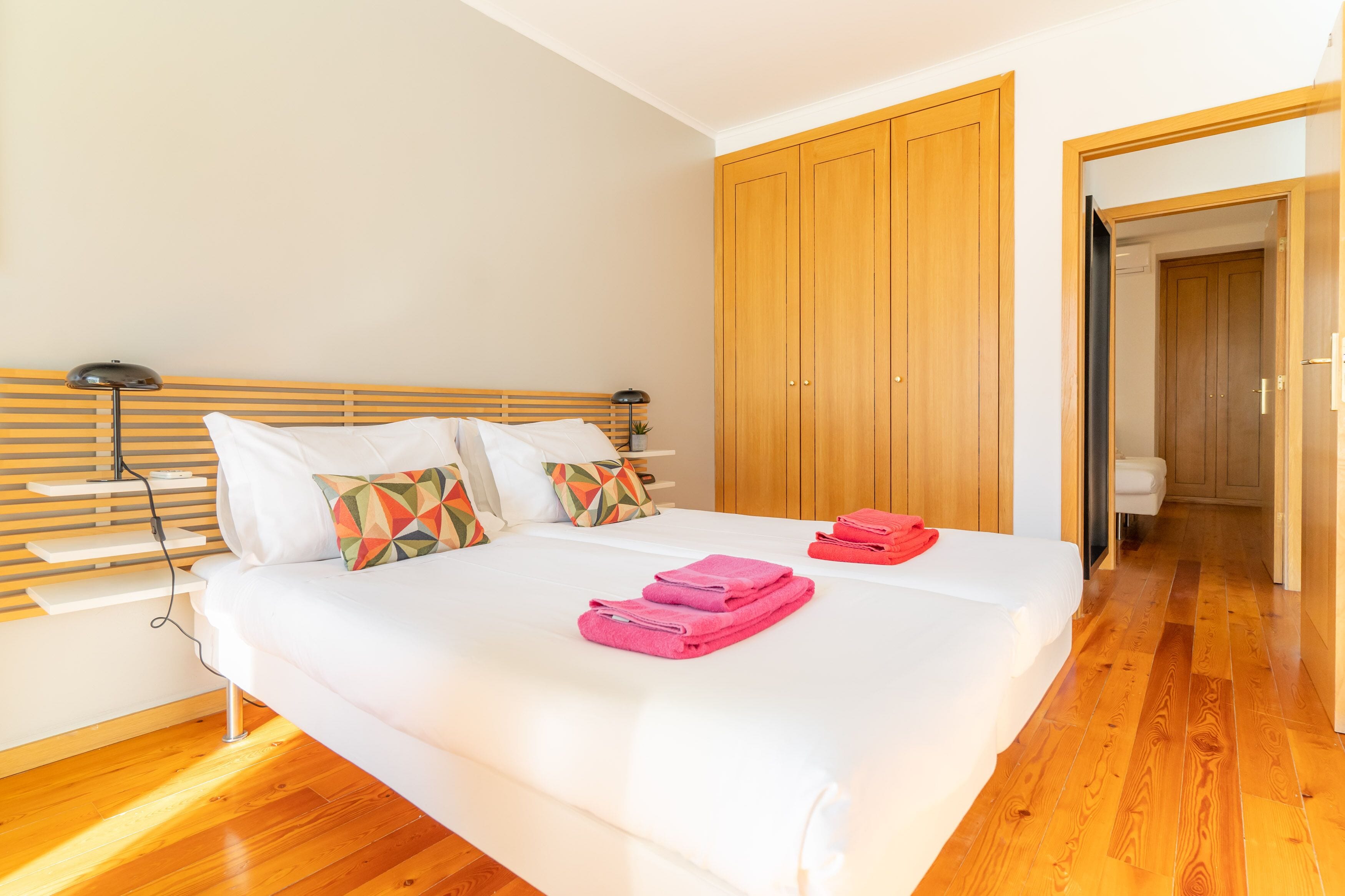 A neatly made bed with white linens and pink towels, a wooden headboard, and a wooden wardrobe in a bright room with wooden flooring.