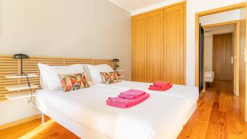 A neatly made bed with white linens and pink towels, a wooden headboard, and a wooden wardrobe in a bright room with wooden flooring.