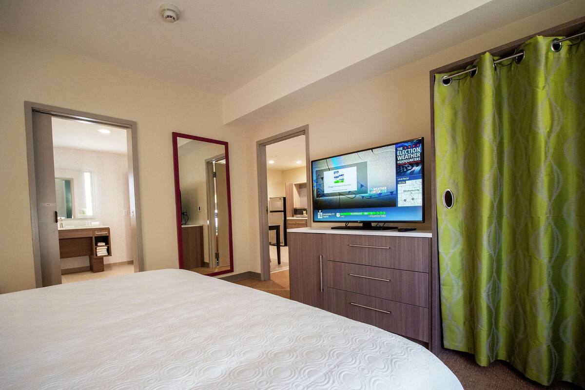 A hotel room with a large bed, a television displaying weather information, a mirror, and a bathroom with a sink and mirror.