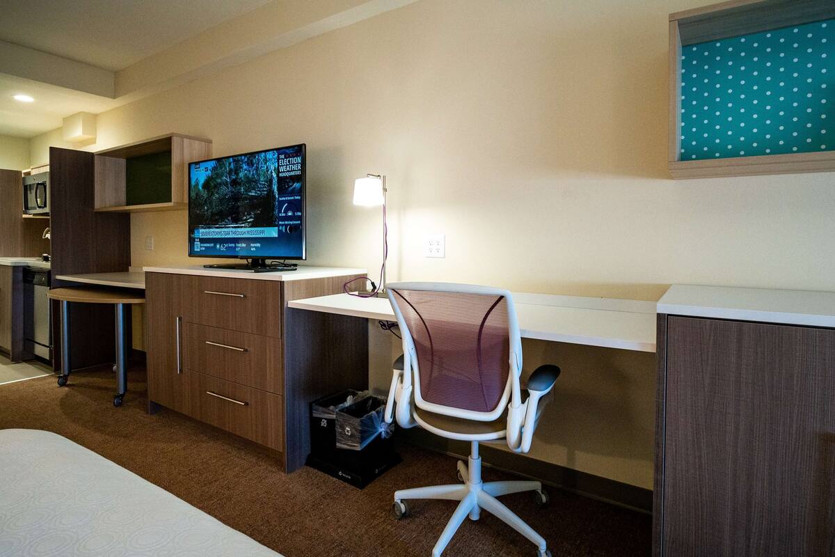 A hotel room with a desk, chair, television, and a bulletin board.