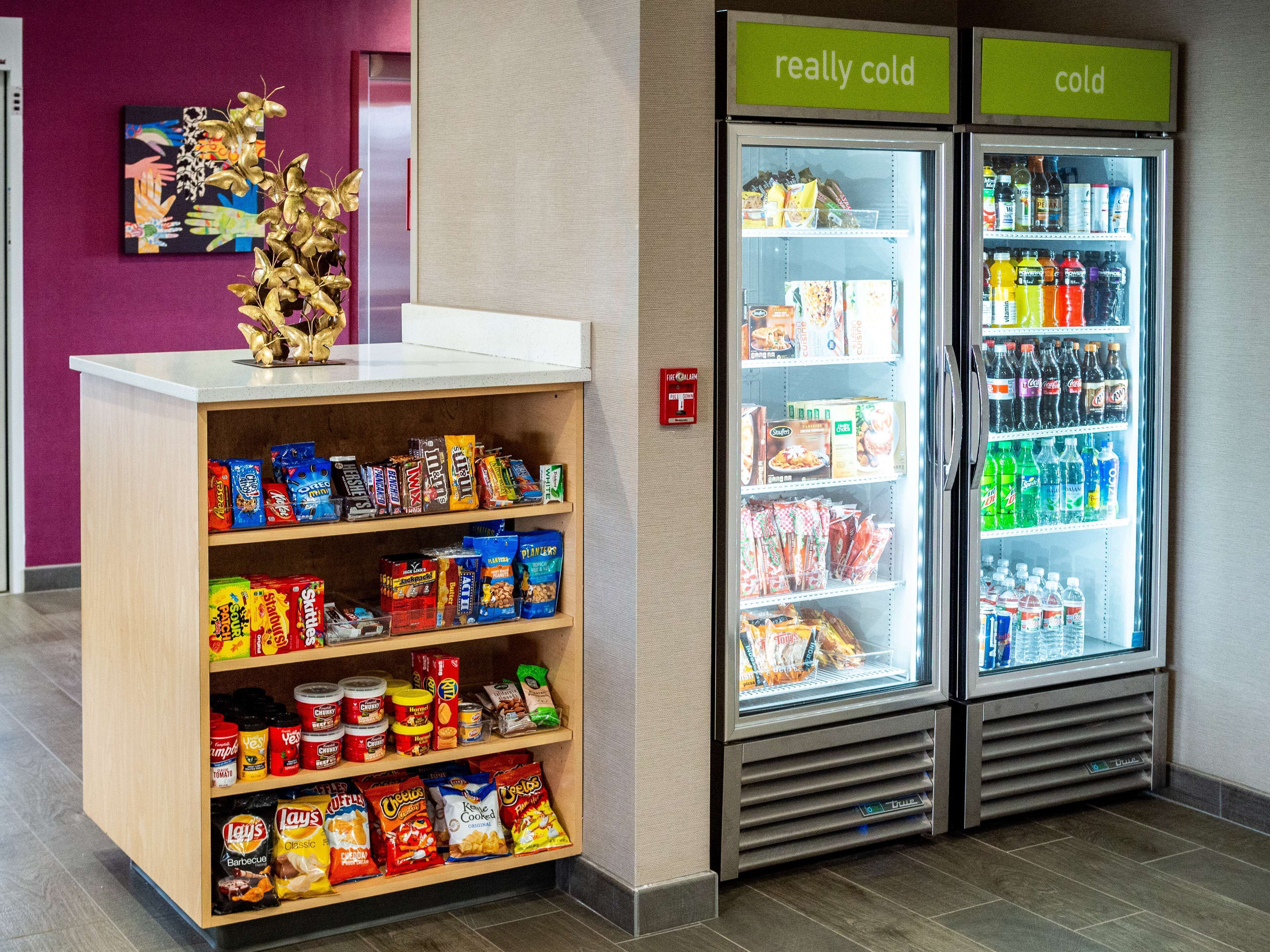 Een hapjesdisplay met verschillende chips en snacks, een koelkast met drankjes en een decoratieve plant.
