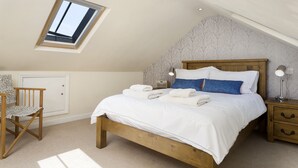 A bedroom with a sloped ceiling, a skylight, a wooden bed, a bedside table with a lamp, and a chair.