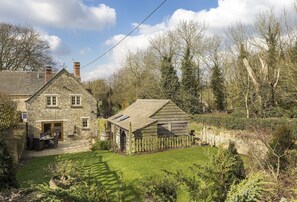 A stone house with a small attached wooden structure, surrounded by a grassy yard and trees.