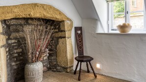 Une pièce avec une cheminée en pierre, un vase contenant des plantes séchées, une chaise en bois et une fenêtre avec un store à carreaux.