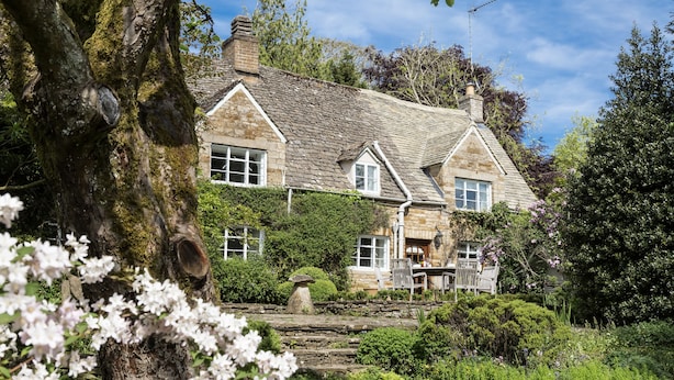 Une maison en pierre avec un toit en pente, entourée d’un jardin où poussent des plantes à fleurs et bordée d’un chemin en pierre.