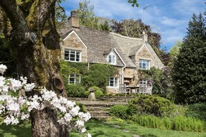 A stone house with a sloped roof, surrounded by a garden with flowering plants and a stone pathway. - Top Cottage, Oddington, Upper and Lower Oddington, Stow-on-the-Wold (Upper Oddington)