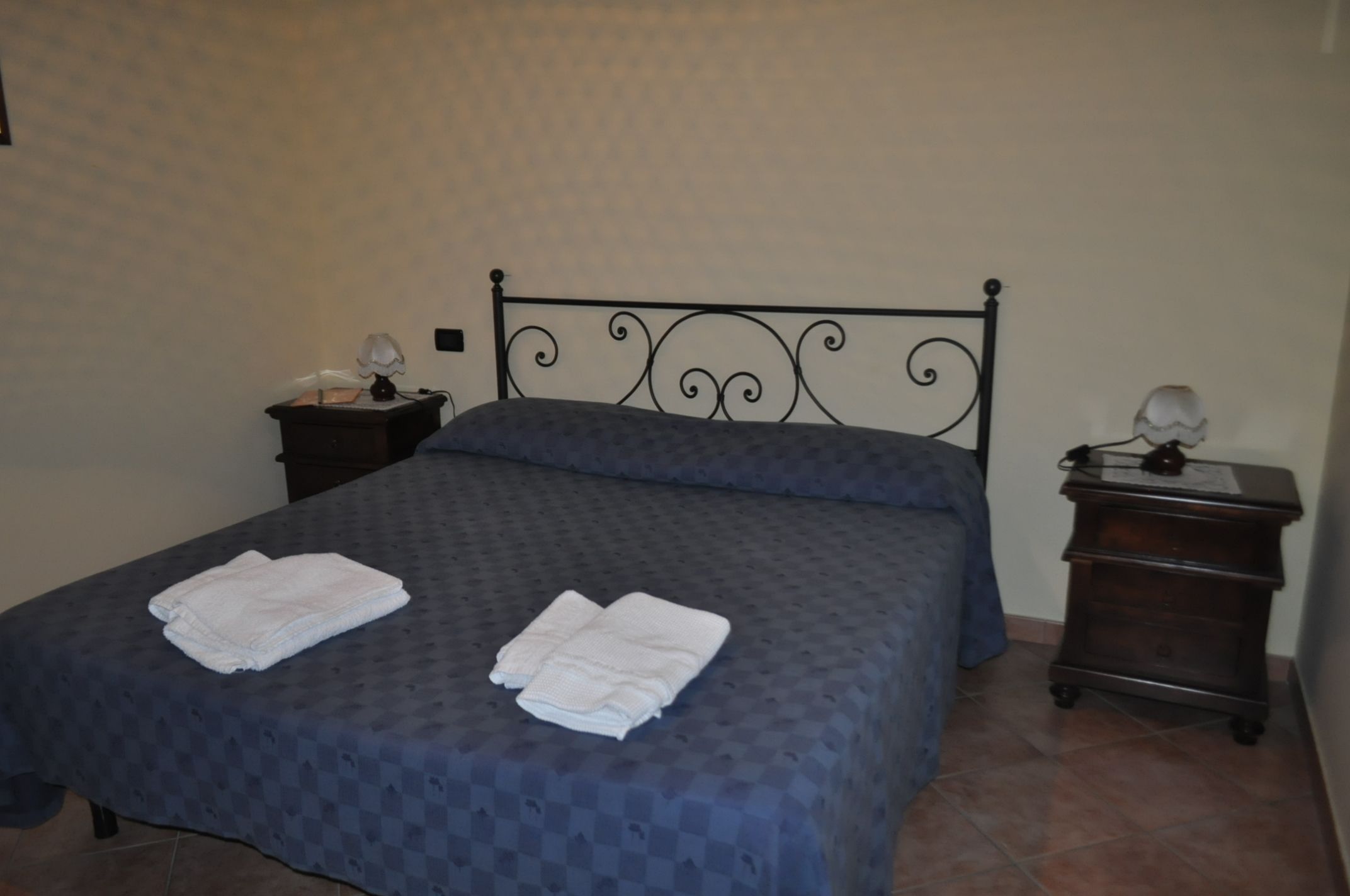 A neatly made bed with a blue bedspread and two folded white towels. There are two bedside tables, each with a lamp.