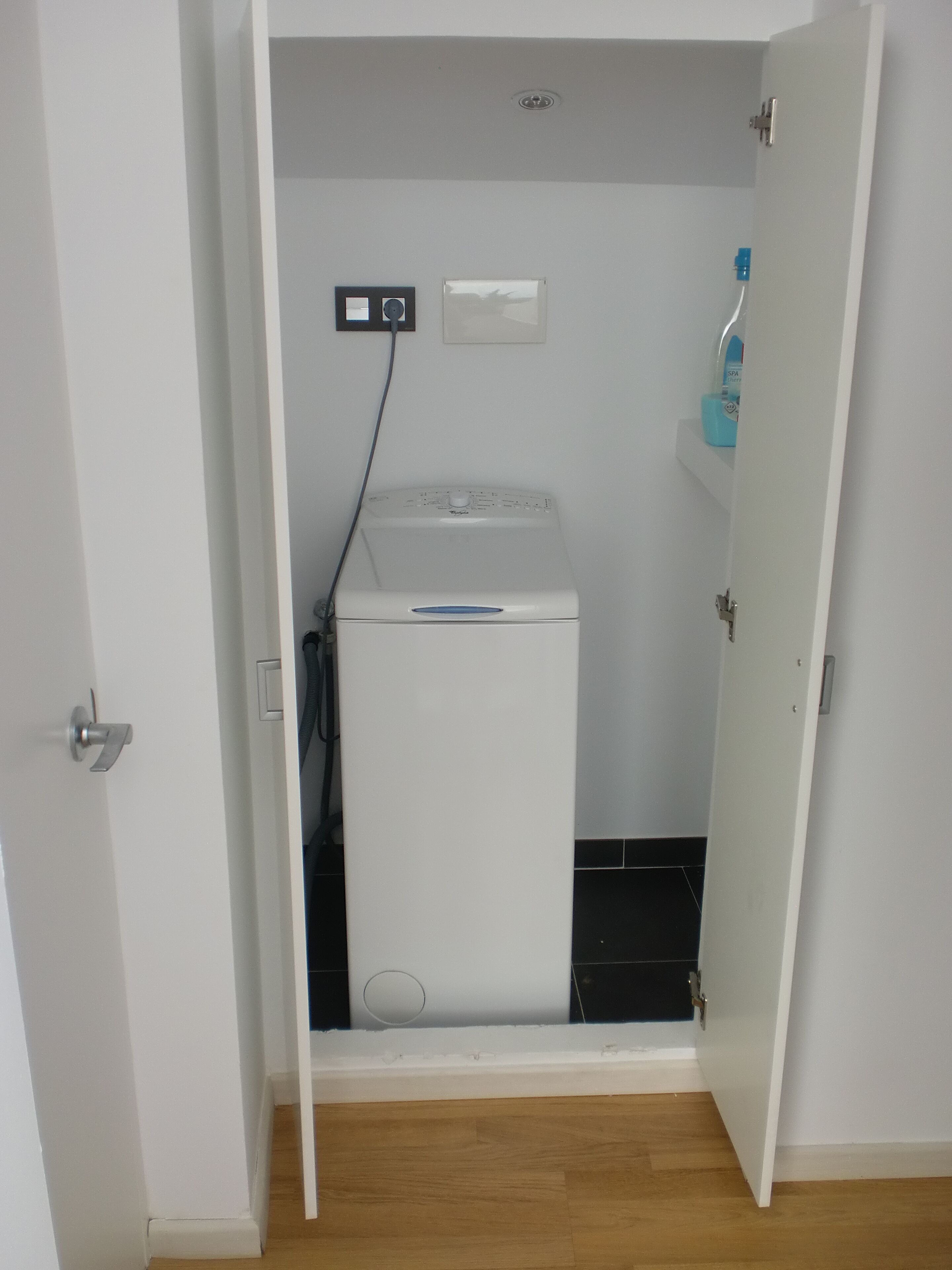 A small laundry room with a washing machine, a power outlet, and a water dispenser on the wall.