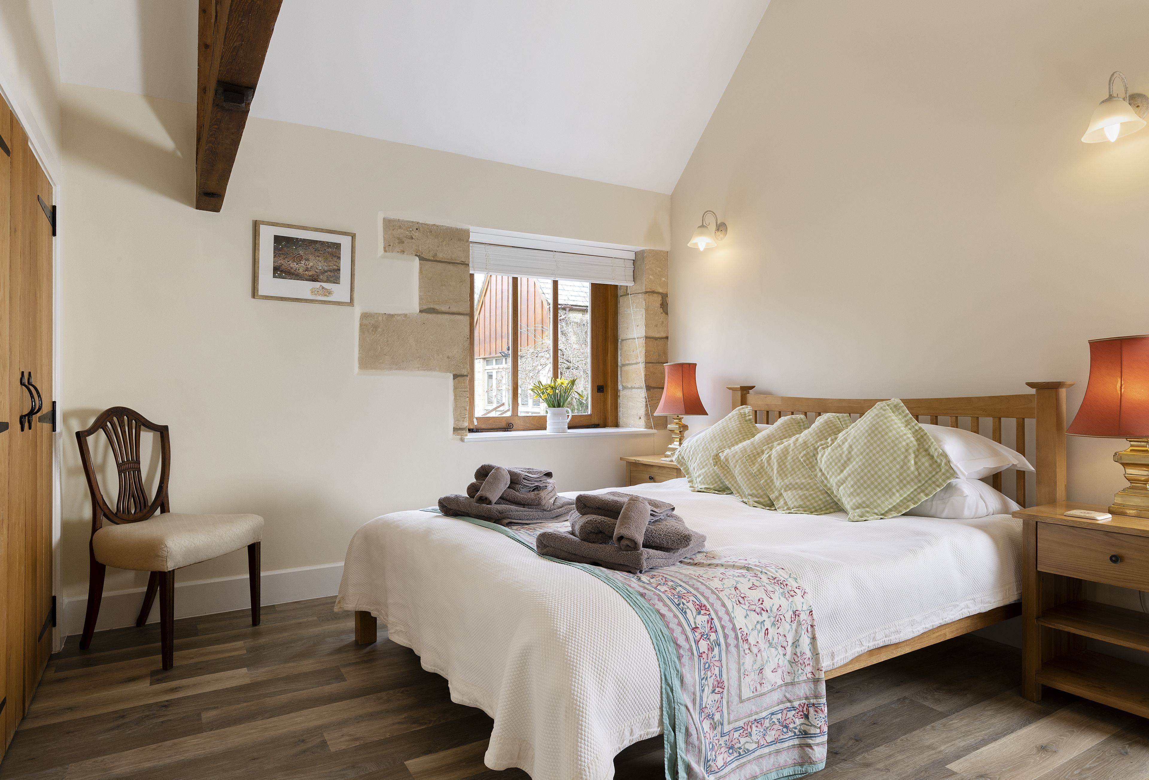 A bedroom with a bed, bedside tables, a chair, a window, and a wooden ceiling beam.