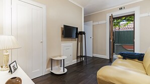 Een kamer met een beige bank, een klein rond tafeltje met een lamp, een aan de muur bevestigde televisie en een deur naar een buitenruimte.