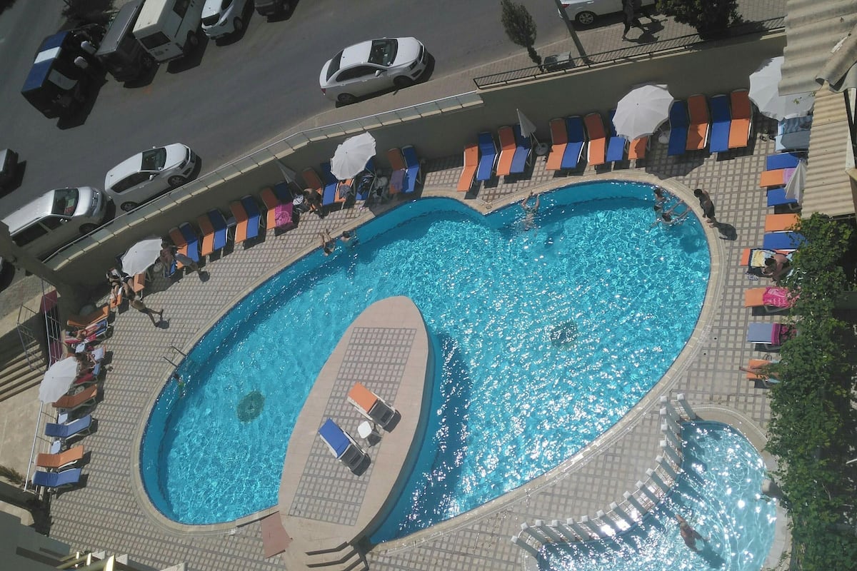 An aerial view of a swimming pool with sun loungers and umbrellas, surrounded by a tiled walkway and adjacent buildings.