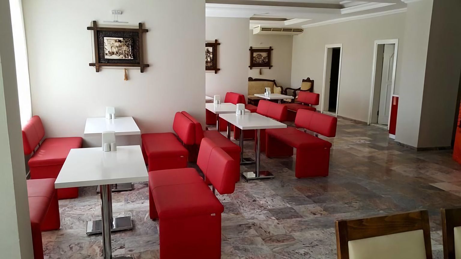 Un espace repas avec des banquettes rembourrées de tissu rouge et des tables blanches, un sol en marbre et des œuvres d’art encadrées accrochées aux murs.