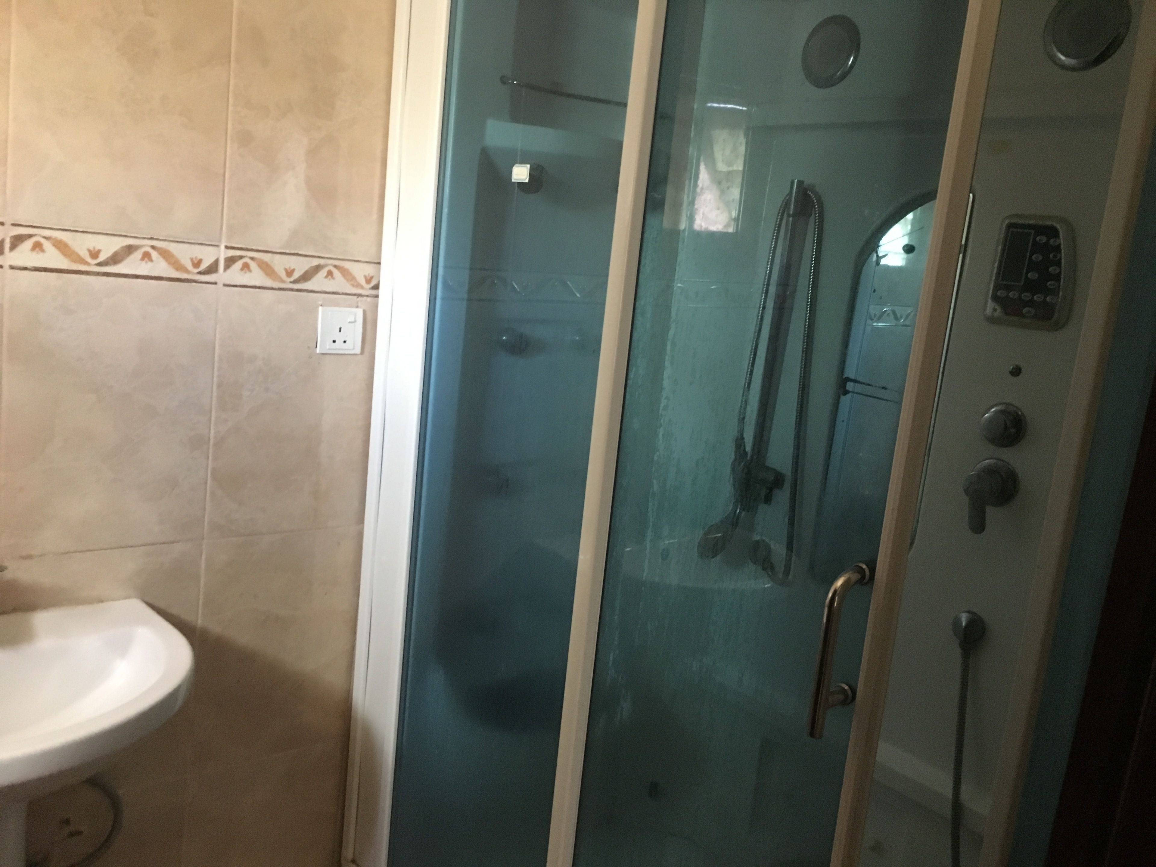 Un box de ducha con paredes de vidrio, un panel de control, un lavamanos blanco y paredes con azulejos.