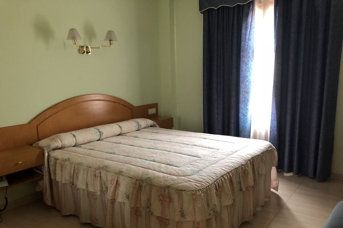 A bedroom with a wooden bed, a nightstand, a lamp, and a window with curtains.