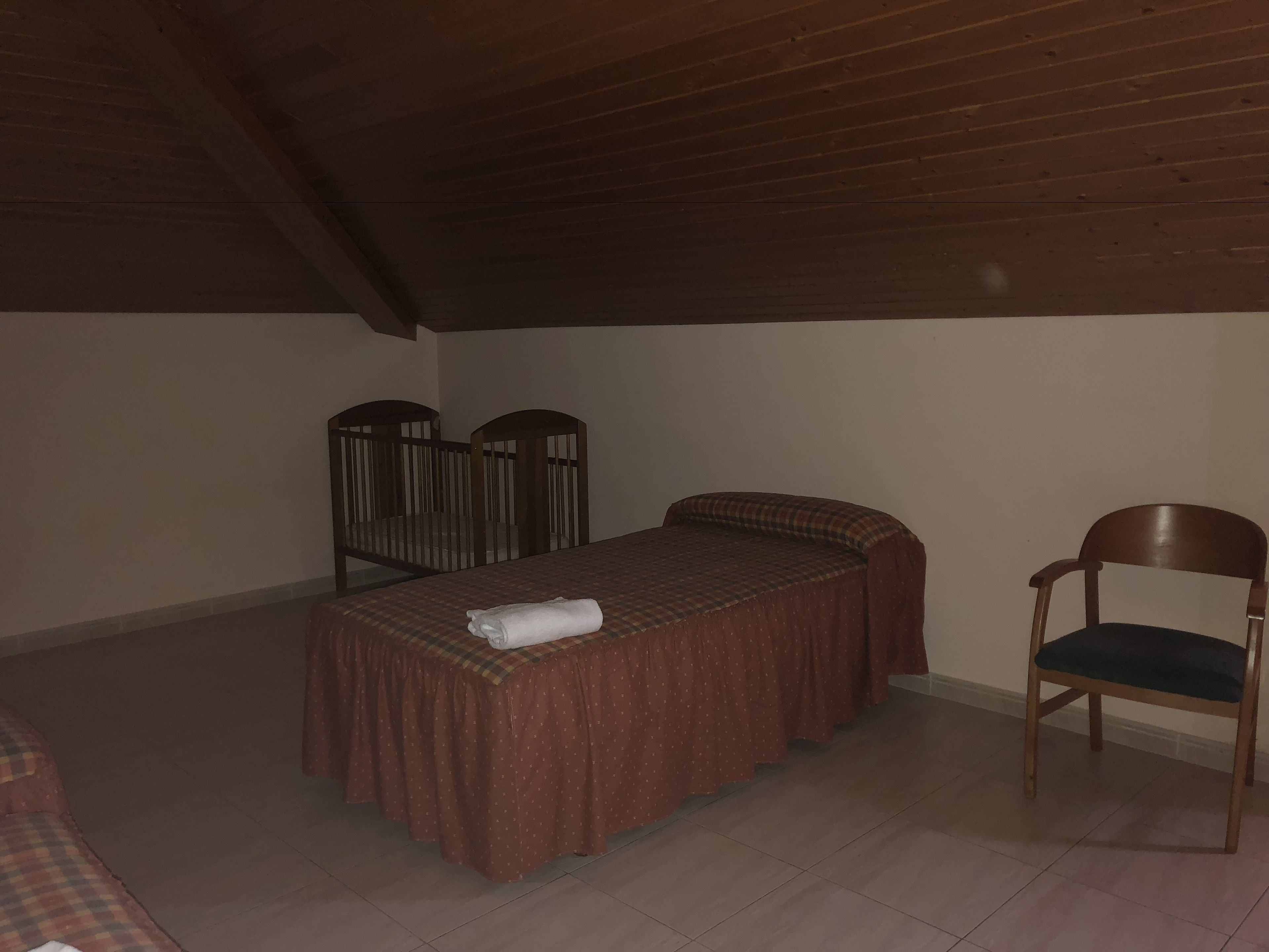 Una habitación pequeña y con poca luz, con una cama individual, una silla y techo de madera.
