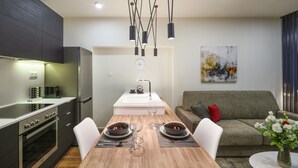 A modern kitchen with a built-in oven, a dining table set for two with a vase of flowers, and a grey sofa in the living area. - Olvios Design Suite (Thessaloniki)