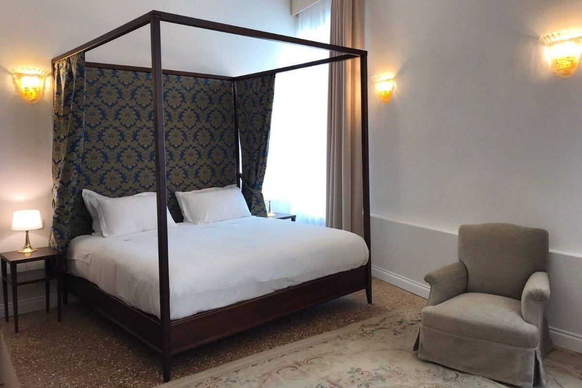 A four-poster bed with a canopy, a bedside table with a lamp, a chair, and a patterned carpet.