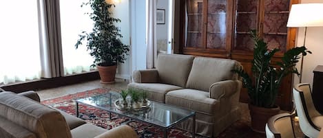 A living room with a beige sofa, a glass coffee table, and a patterned rug. There is a large potted plant and a cabinet with glass doors.