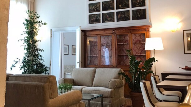 A cozy living room with a high ceiling, a fireplace, a large window with curtains, and a collection of framed pictures on the wall. - Suites alla Maddalena (Venice)