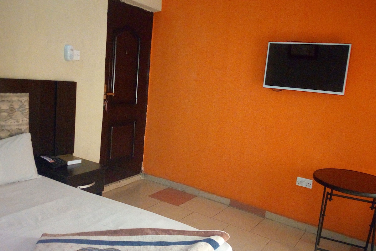A hotel room with a bed, a TV mounted on the orange wall, a small table, and a door.