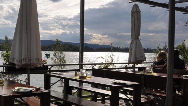 Een terras met houten tafels en banken, parasols en uitzicht op een meer en bergen.