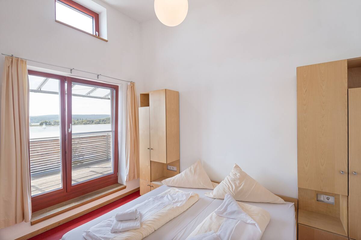 A bedroom with a bed, a wooden wardrobe, and a window with a view of a body of water.