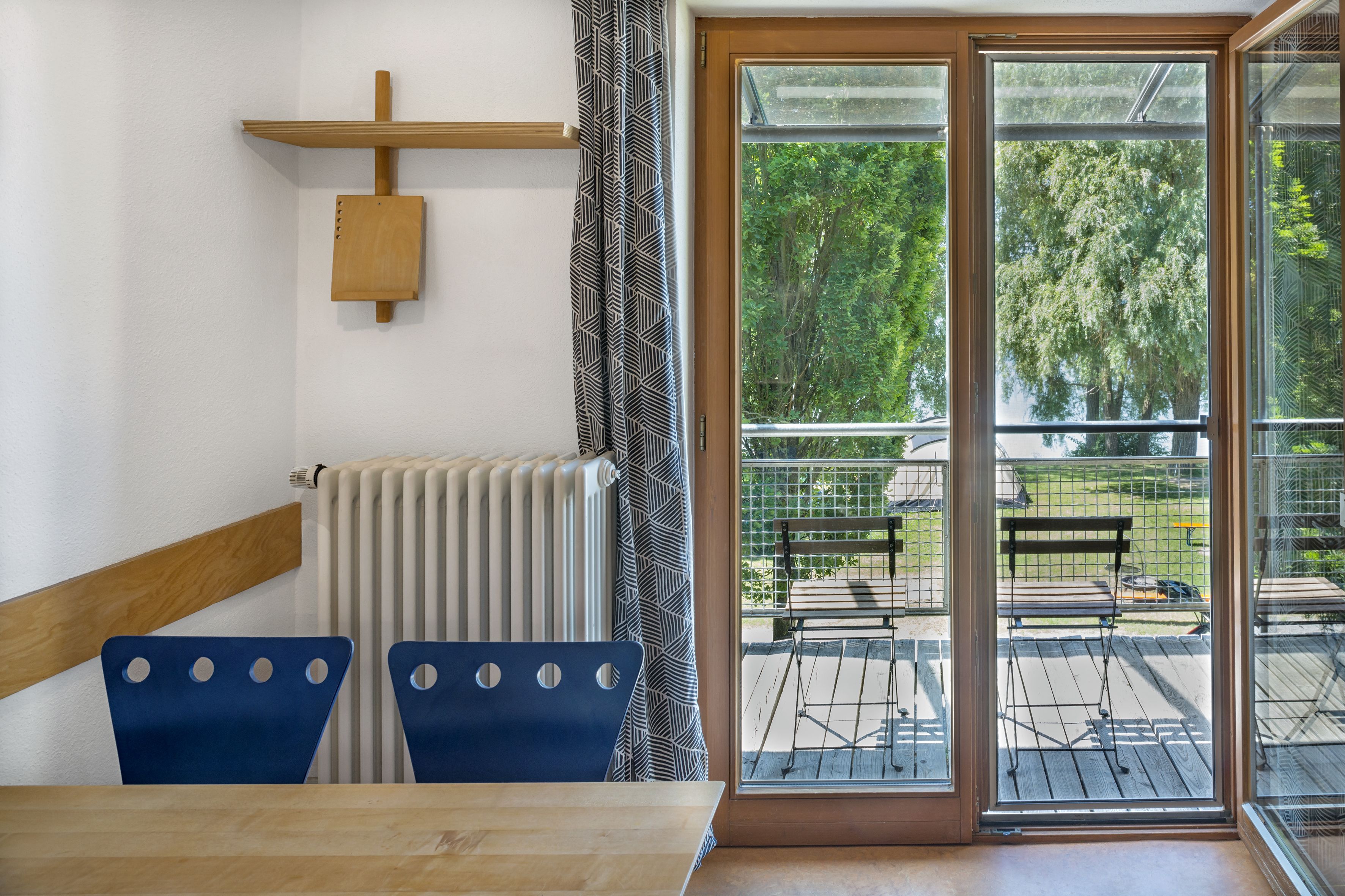 A room with a wooden table and two blue chairs, a radiator, a wooden shelf, and a sliding glass door leading to an outdoor patio with a bench.
