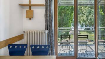 Una habitación con una mesa de madera y dos sillas azules, un radiador, un estante de madera y un ventanal corredero que da a un patio exterior con una banca.