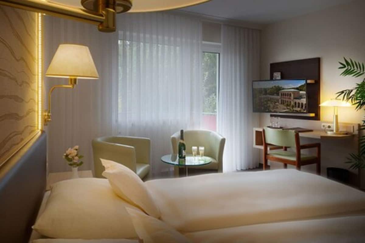 A hotel room with a large bed, two armchairs, a small round table with a bottle and glasses, a desk with a chair, a TV mounted on the wall, and a plant.