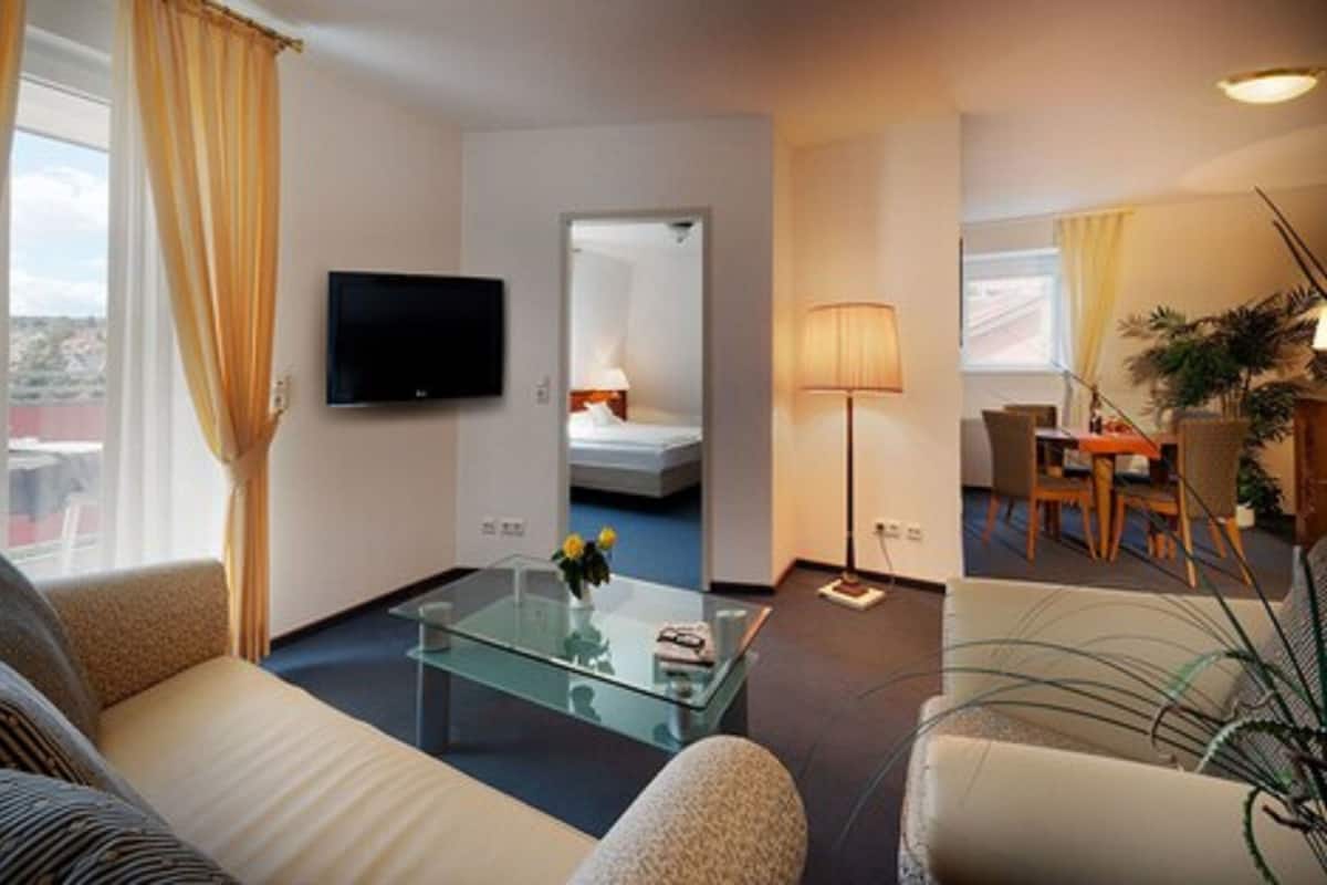 A hotel room with a sofa, a glass coffee table, a TV, a bed, a desk, and a chair.