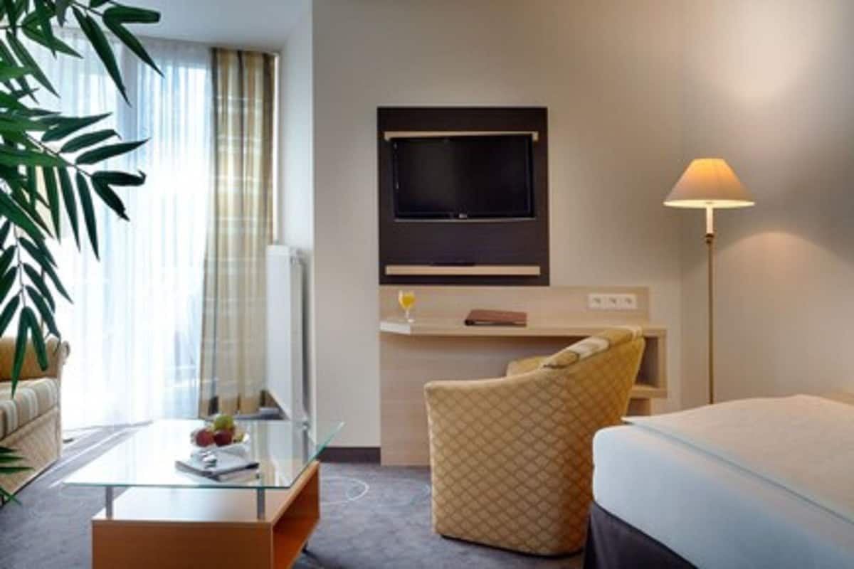 A hotel room with a bed, a glass coffee table, a sofa, a TV mounted on the wall, and a floor lamp.