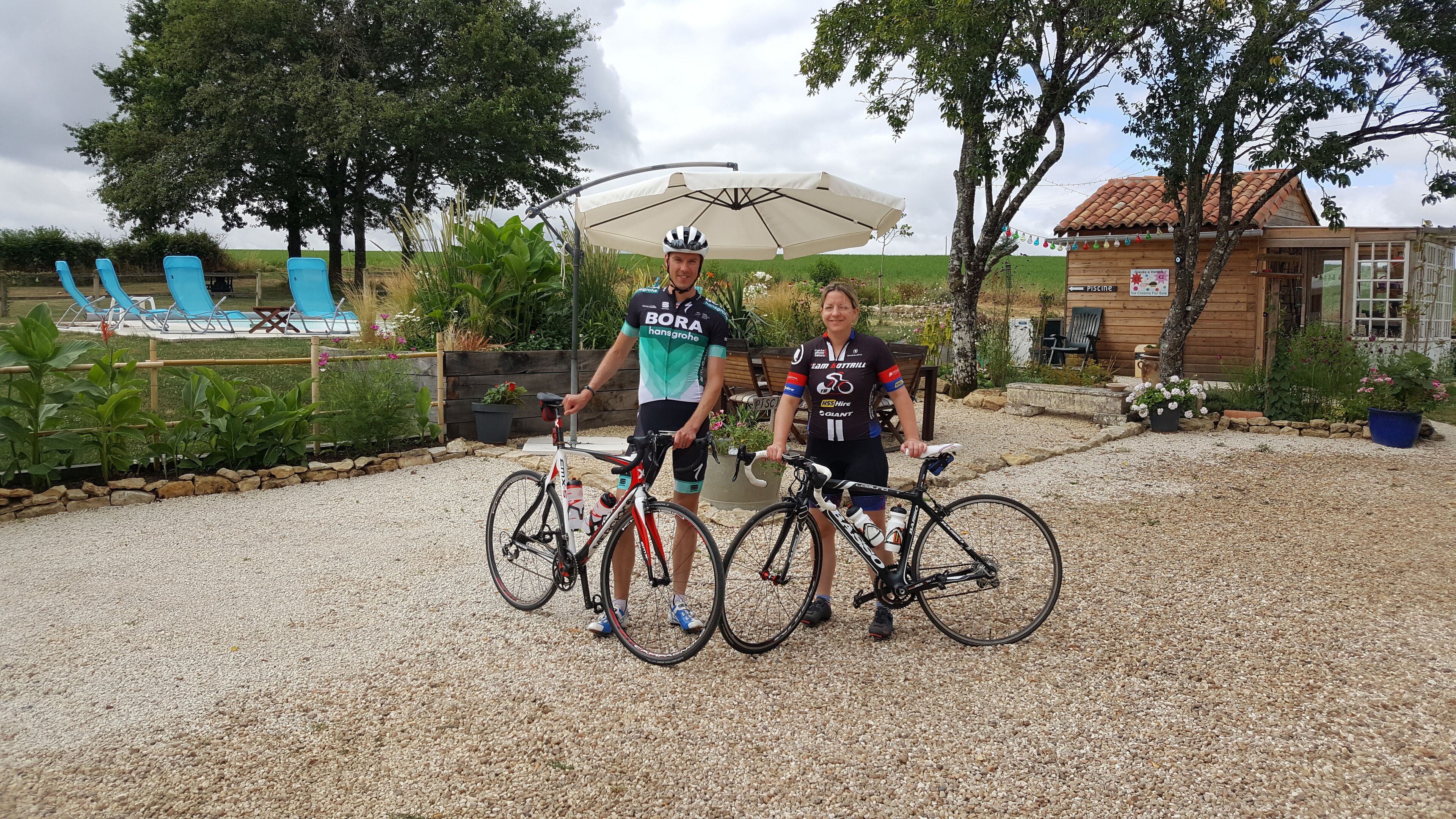 Two people standing next to bicycles in a gravel area with a pool, lounge chairs, and a wooden building in the background.