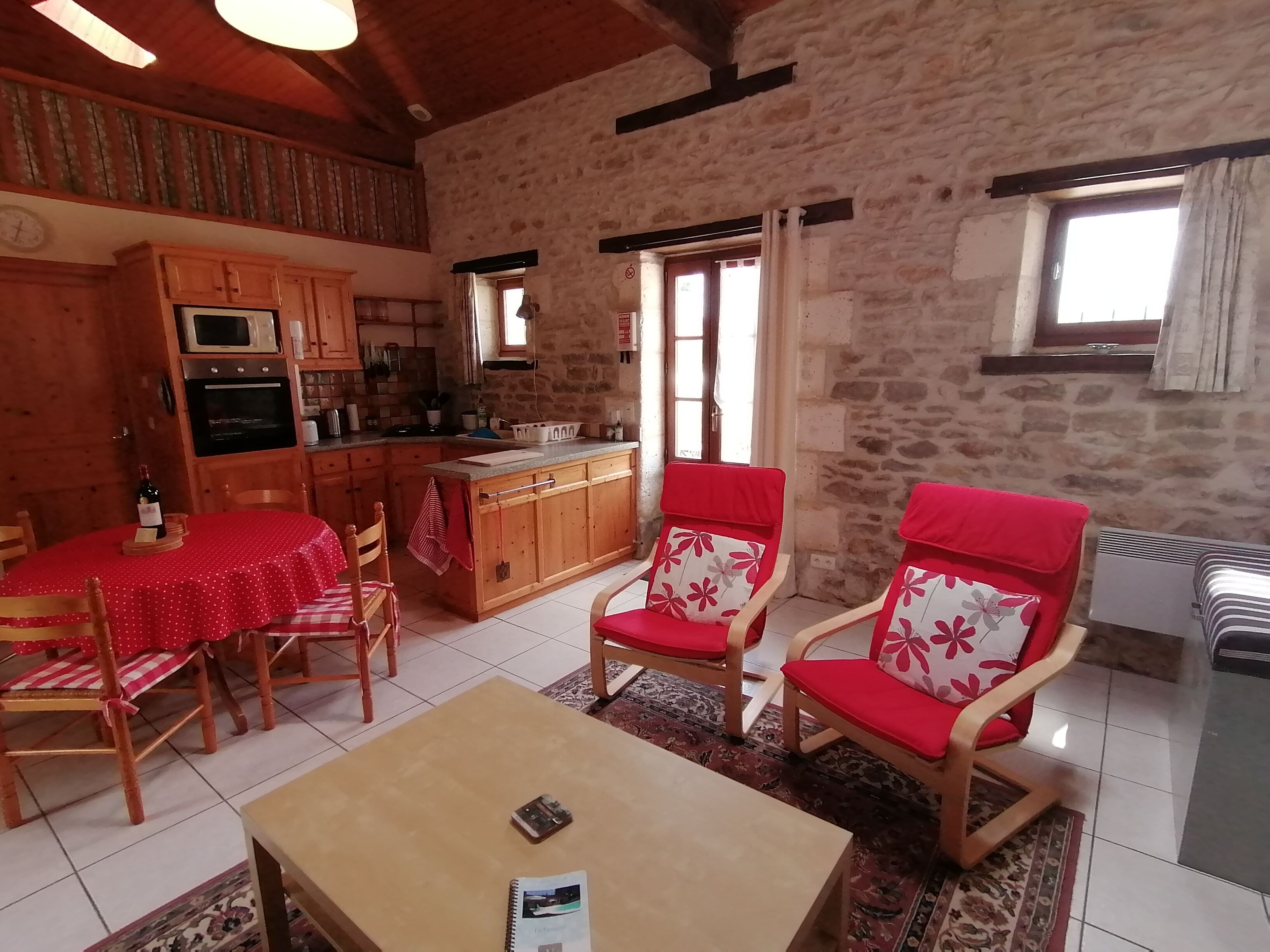 A cozy living room with a dining area, featuring wooden furniture, a red and white patterned sofa, and a kitchen with stone walls and wooden beams.
