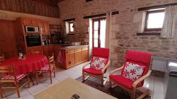 A cozy living room with a dining area, featuring wooden furniture, a red and white patterned sofa, and a kitchen with stone walls and wooden beams.