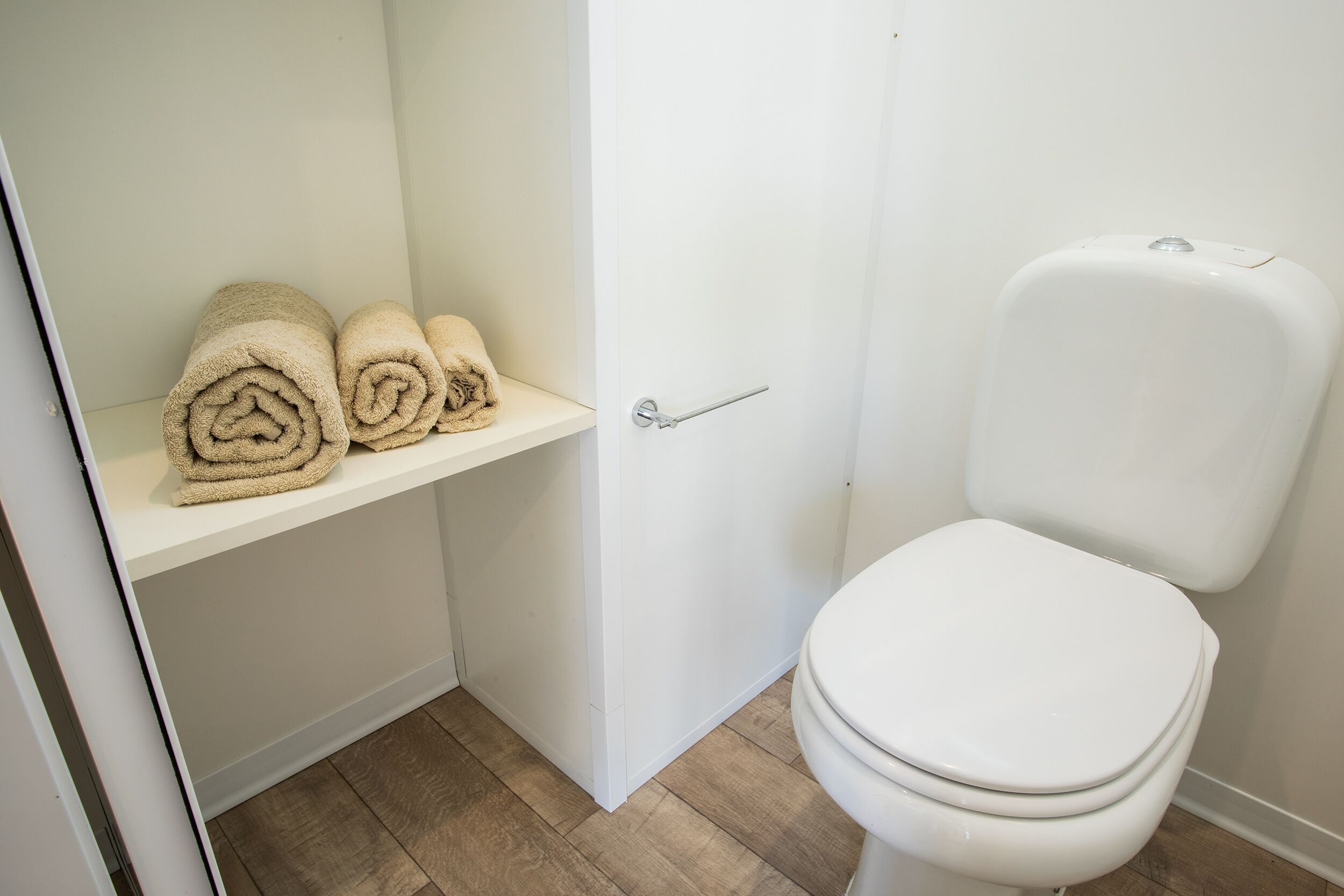 Une salle de bain avec des toilettes, une étagère où sont pliés des serviettes et un plancher en bois.