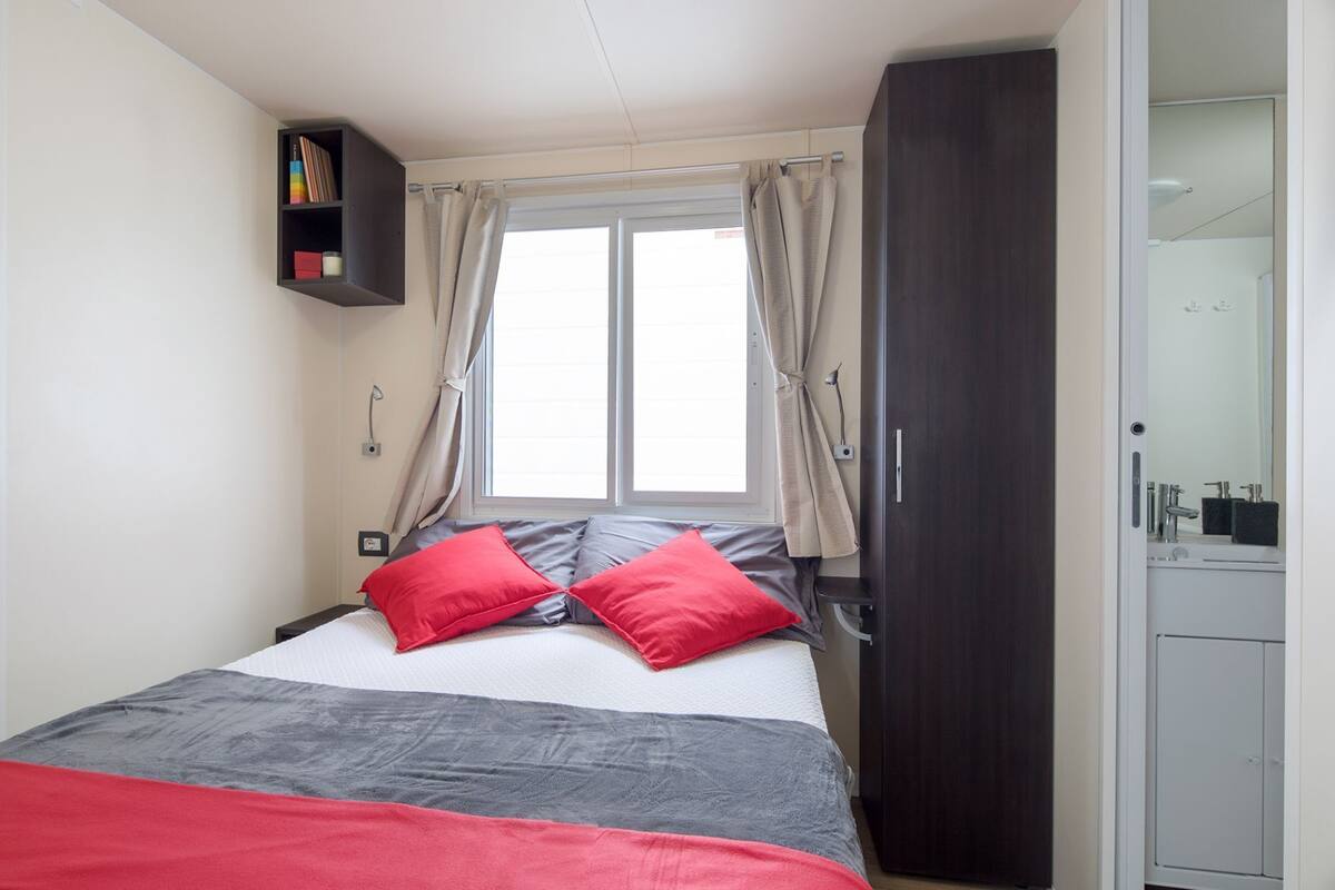 A bedroom with a bed, two red pillows, a grey blanket, a window with curtains, a wardrobe, and a small shelf with books.
