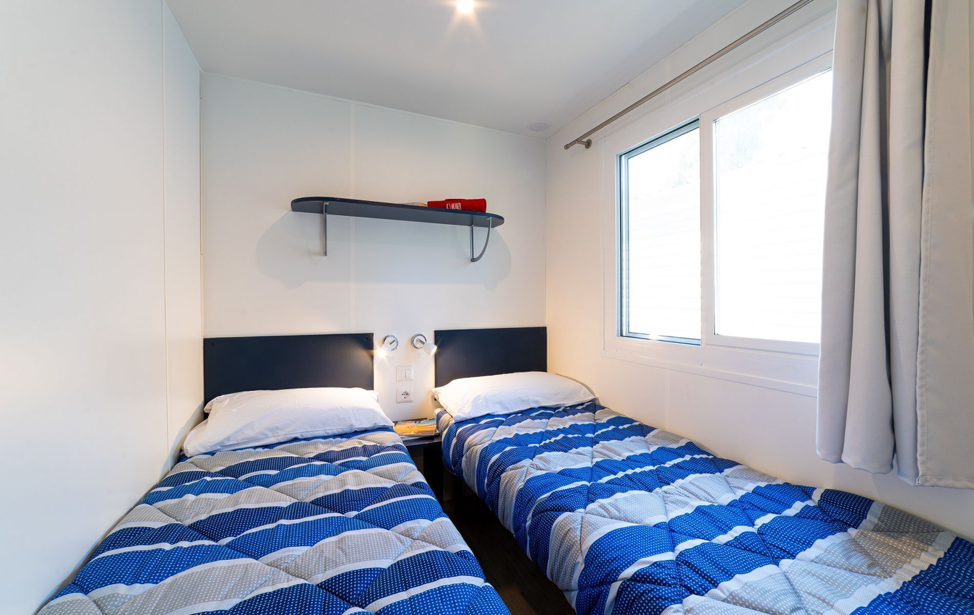 A small, well-lit room with two single beds, a shelf above the beds, and a window on one side.