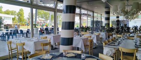 A restaurant with outdoor seating, large windows, and a view of a playground. Tables are set with white tablecloths and chairs.